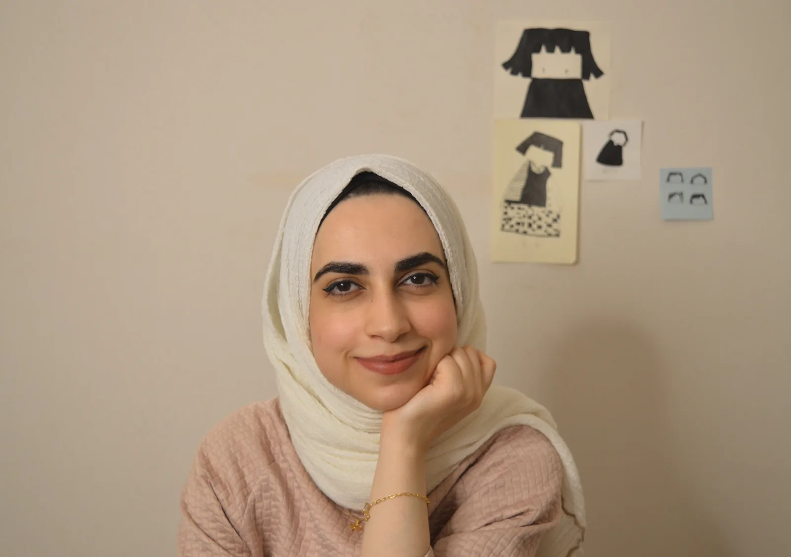Rama Aldakkak resting her chin on her hand, her illustrations pinned to the wall behind her